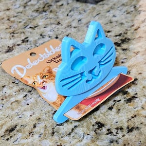 May include: A light blue cat-shaped treat holder sits atop a package of cat treats. The treat holder is made of plastic and has a cutout design of a cat's face. The treat package features a picture of a cat and the word "Delectables".