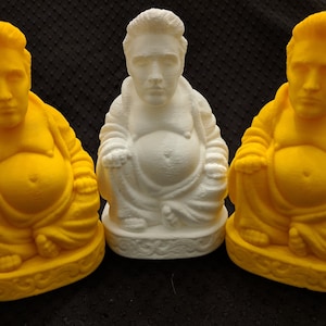 May include: Three ceramic figurines of a person in a seated meditation pose. The figurines are painted in yellow and white. The middle figurine is white, while the figurines on either side are yellow.