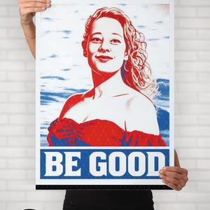 May include: Poster with a red and blue graphic of a woman with the words "BE GOOD" in bold blue letters. The poster is held up by a person in front of a white brick wall. The website "woke-wear.com" is visible.