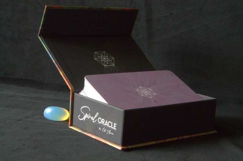 Może przedstawiać: Zamknięte pudełko z fioletowym zestawem kart w środku. Pudełko jest czarne z kolorowym wzorem na spodzie i tekstem "Spiral Oracle by Liz A."