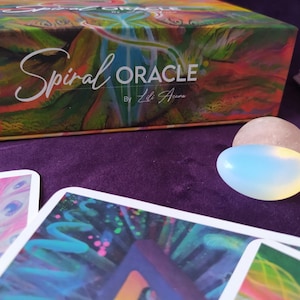 Może przedstawiać: Pudełko na talię kart "Spiral Oracle" autorstwa Lili Auna. Pudełko jest kolorowe z abstrakcyjnym wzorem w kształcie spirali. Pudełko leży na fioletowym tle z dwoma gładkimi, owalnymi kamieniami.