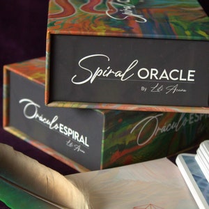 Może przedstawiać: Dwa pudełka z kolorową abstrakcyjną sztuką z przodu. G&oacute;rne pudełko ma czarną etykietę z białym tekstem, kt&oacute;ry brzmi "Spiral Oracle by Lili Acs.". Dolne pudełko ma czarną etykietę z białym tekstem, kt&oacute;ry brzmi "Oraculo Espiral Lili Acs."