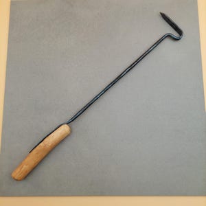 Puede incluir: Una herramienta de metal negro con mango de madera. La herramienta tiene un eje largo y delgado con un extremo curvado y una punta. El mango es de madera de color claro.