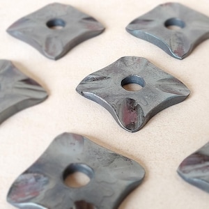 Op de afbeelding: Close-up van verschillende grijze, ruitvormige metalen ringen met een centraal gat. De ringen hebben een getextureerd oppervlak en afgeronde randen. Het metaal heeft een licht verouderd uiterlijk met hints van roodbruin. De ringen zijn gerangschikt op een lichtgekleurd oppervlak.