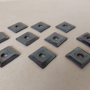 Puede incluir: Diez piezas cuadradas de ferretería metálica con un agujero central. Las piezas tienen un tono gris verdoso oscuro con bordes marrón rojizo. La ferretería está dispuesta sobre una superficie de color beige claro.