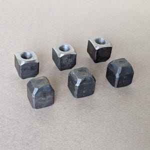 May include: A collection of six dark grey metal square nuts with bevelled edges. Each nut features a central threaded hole, showcasing a raw, industrial finish with visible metallic textures and distinct geometric shapes arranged in a cluster.