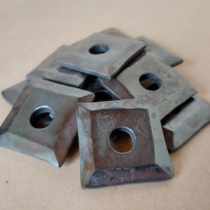 Puede incluir: Un montón de insertos metálicos cuadrados con un agujero central. Los insertos tienen un aspecto oscuro y envejecido con desgaste visible. Los bordes están ligeramente biselados, lo que sugiere un tema vintage o industrial.