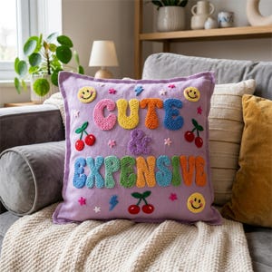 Op de afbeelding: Een vierkant lavendelkleurig sierkussen met veelkleurig punch needle borduurwerk. De tekst luidt CUTE & EXPENSIVE in dikke, getextureerde letters. Decoratieve motieven zijn onder meer gele smileys, rode kersen en kleine blauwe bliksemschichten.