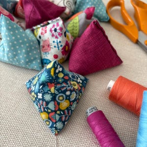 Puede incluir: Una colección de alfileteros de tela con forma de pirámide en varios patrones coloridos, incluyendo flores, lunares y estampados de gatos. Se ven tijeras naranjas, un carrete de hilo naranja y un carrete de hilo magenta con la etiqueta 282.
