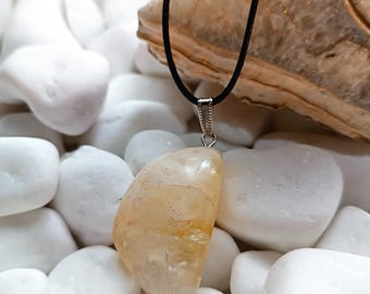 Puede incluir: Un collar con colgante que presenta una piedra preciosa pulida, translúcida, de color naranja claro. La piedra está suspendida de un cordón negro con un cierre de metal plateado. El collar se exhibe sobre un lecho de piedras blancas.
