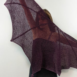 May include: A deep purple knitted shawl with a delicate lace pattern and a tassel. The shawl is draped over a person's shoulders, with the person's head and face obscured by the shawl.