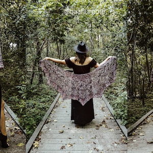 May include: A woman wearing a black dress and a black hat is standing on a wooden walkway in a forest. She is holding a purple and gray shawl in front of her. The shawl is draped over her shoulders and is flowing in the wind.