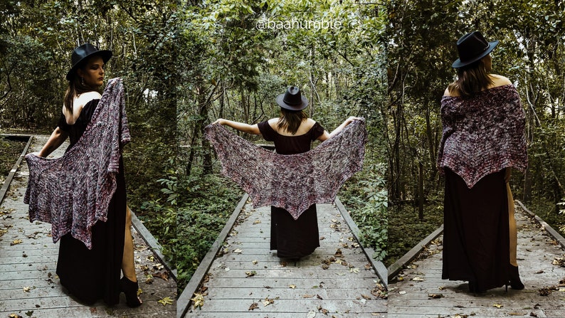 May include: A woman wearing a black dress and a black hat is standing on a wooden walkway in a forest. She is holding a purple and gray shawl in front of her. The shawl is draped over her shoulders and is flowing in the wind.