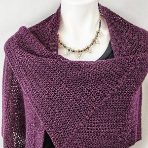 May include: A purple knitted shawl draped over a white mannequin. The shawl has a delicate openwork pattern and a subtle sheen. A gold necklace with triangular pendants is worn over the shawl.