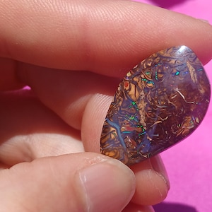 Pode incluir: Uma pedra preciosa de opala polida em forma de gota. A pedra exibe um padrão complexo de tons marrons, dourados e azuis com flashes iridescentes de verde e laranja. A opala é segurada entre os dedos contra um fundo rosa.