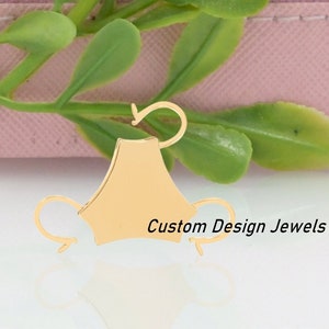 May include: Gold-toned metal earring with a unique design featuring a curved shape and a small loop. The earring is on a pink surface with green foliage.