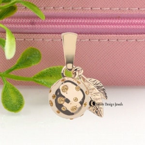 May include: A gold charm pendant in the shape of a small orange with leaves. The orange has a textured surface with small gold dots. The pendant is on a pink background.