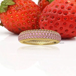 Pode incluir: Um anel de banda de ouro com quatro fileiras de pedras preciosas cor-de-rosa. O anel está colocado sobre um fundo de morangos vermelhos.