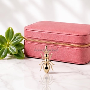 May include: A gold-toned ant-shaped pendant featuring a small black circular accent on its abdomen. The pendant is positioned in front of a rectangular pink jewellery box with a gold zip and a small green succulent plant to the left side.