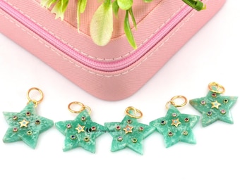 May include: Five star-shaped pendants with gold loops. The pendants are light green and decorated with small, colourful gemstones. A pink case and green plant sprig are in the background.