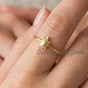 May include: A gold-toned ring featuring a small, detailed turtle charm centered on a thin, polished band. The ring is worn on a finger, showcasing the delicate metalwork and the subtle, embossed text that reads Custom Name Jewels on the band.