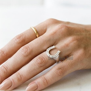 May include: A hand adorned with a gold band ring and a silver horseshoe ring set with small, clear stones. A delicate silver bracelet with small stones is also visible. The jewellery is set against a light background.