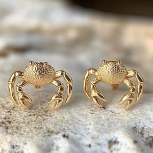 May include: A pair of gold-toned crab-shaped stud earrings. The earrings are small and delicate, with a textured surface.