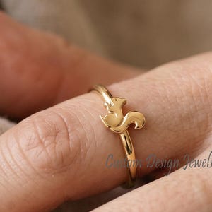 May include: A gold-toned ring featuring a small, stylized squirrel figure as the central focal point. The band is thin and polished, showcasing a smooth metallic finish. The squirrel design is integrated directly into the top of the ring band.