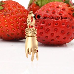May include: A gold charm in the shape of a hand with a red gemstone on the ring finger. The charm is sitting on top of three red strawberries.
