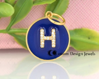Puede incluir: Encanto de oro con un círculo de esmalte azul y una letra H de cristal transparente. El encanto está sobre una superficie blanca con un reflejo.