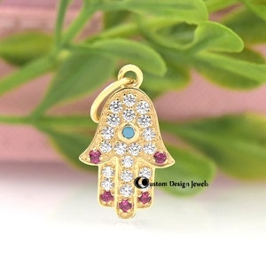 May include: Gold charm in the shape of a hamsa hand with a blue evil eye and red gemstones. The charm is set with clear cubic zirconia stones.