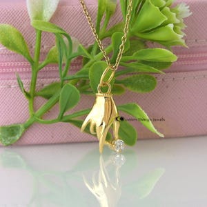 Op de afbeelding: Een gouden ketting met een hanger in de vorm van een hand die een kleine diamant vasthoudt. De hanger hangt aan een delicate gouden ketting.