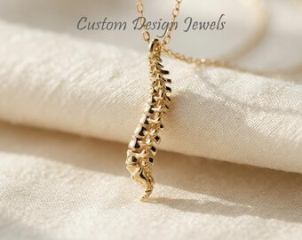 May include: A gold-toned necklace featuring a pendant shaped like a human spinal column. The pendant is suspended from a delicate gold-toned chain. The text Custom Design Jewels is visible in the upper portion of the image, positioned above the pendant.