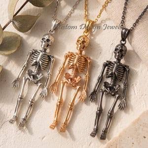 May include: Three skeleton pendants on chains in silver, gold, and dark gunmetal tones. Each pendant features a detailed human skeleton figure with articulated limbs. The text Custom Design Jewels is visible in the center.