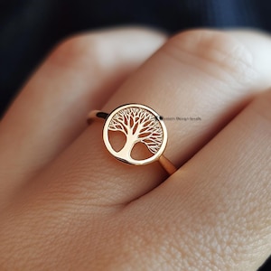 May include: A gold ring featuring a tree of life design. The ring is worn on a finger.