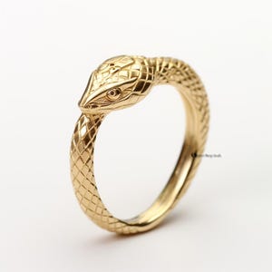 Peut inclure: Une bague dorée en forme de serpent. La tête du serpent est ornée d'écailles et d'un œil, et le corps se courbe pour former l'anneau. La bague est posée sur un fond blanc.