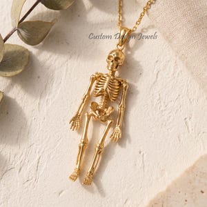 May include: A gold-toned necklace featuring a detailed human skeleton pendant. The pendant is suspended from a delicate gold chain. The text Custom Design Jewels is visible near the top of the skeleton figure, which displays a full anatomical structure.