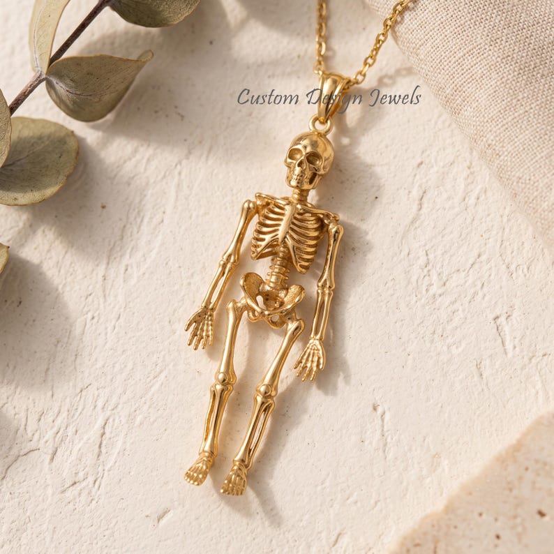 May include: A gold-toned necklace featuring a detailed human skeleton pendant. The pendant is suspended from a delicate gold chain. The text Custom Design Jewels is visible near the top of the skeleton figure, which displays a full anatomical structure.