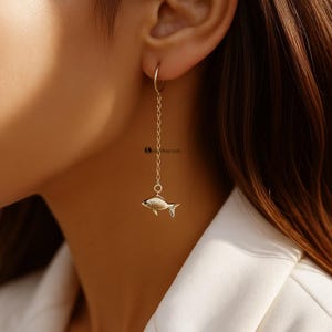 May include: Gold-toned fish earrings. The earrings feature a small fish pendant suspended from a delicate chain, attached to a hoop earring. The fish has detailed scales and fins. The earrings are worn on a person's ear.