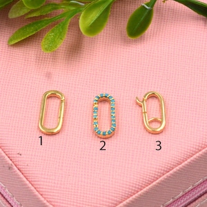 May include: Three gold-colored oval clasps. One is plain, one is adorned with small turquoise-colored stones, and the third has a clasp closure. The clasps are displayed on a pink surface with green foliage.