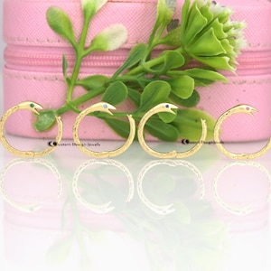 May include: Four gold snake hoop earrings with different colored gemstones in the eyes. The earrings are on a pink surface with a reflection of the earrings below.