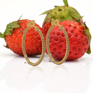 May include: A pair of gold hoop earrings with a row of small diamonds around the edge. The earrings are on a white background with two red strawberries.