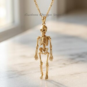 May include: A gold-toned necklace featuring a detailed human skeleton pendant suspended from a matching chain. The pendant displays a full anatomical skeletal structure. The text Custom Design Jewels is visible in the background area.