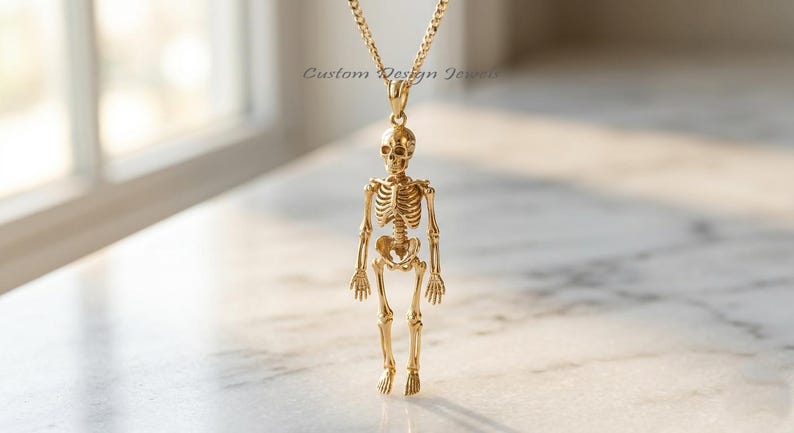 May include: A gold-toned necklace featuring a detailed human skeleton pendant suspended from a matching chain. The pendant displays a full anatomical skeletal structure. The text Custom Design Jewels is visible in the background area.