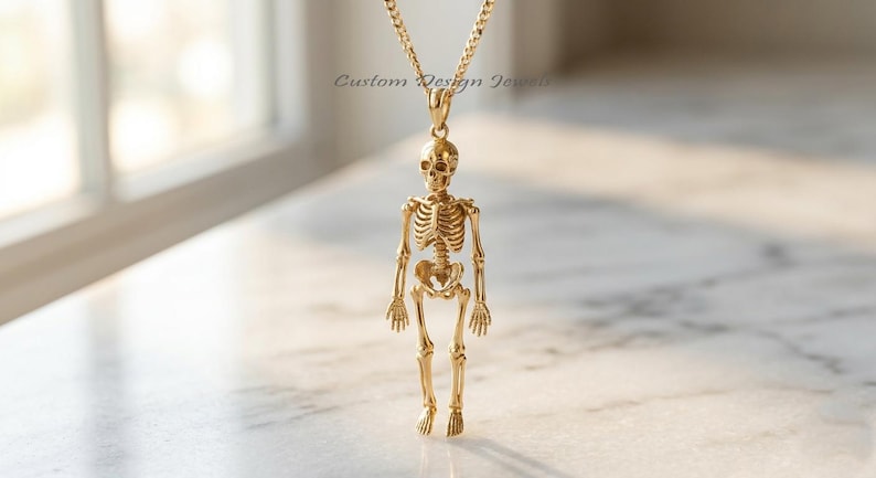 May include: A gold-toned necklace featuring a detailed human skeleton pendant suspended from a matching chain. The pendant displays a full anatomical skeletal structure. The text Custom Design Jewels is visible in the background area.