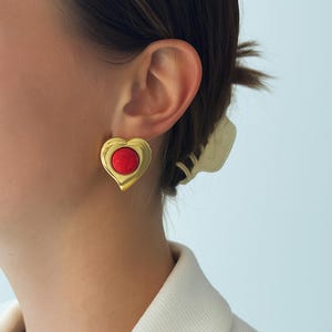 May include: Gold-toned heart-shaped earrings with a vibrant red center stone. The earrings have a smooth, polished finish and a classic design. These earrings are a stylish accessory for any outfit.