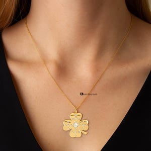 May include: A gold-colored necklace featuring a four-leaf clover pendant. The clover is adorned with small, clear gemstones and a larger gemstone at the center. The necklace is displayed against a dark background.