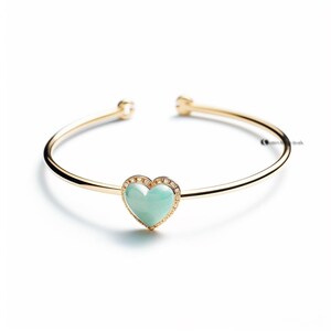 Puede incluir: Pulsera abierta dorada con un colgante en forma de corazón. El colgante presenta una piedra verde claro rodeada por un halo de pequeñas piedras preciosas transparentes. La pulsera tiene un acabado pulido y un diseño simple y elegante.