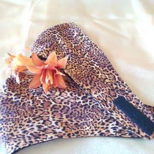 May include: A brown and black leopard print face mask with an adjustable strap and a black hook and loop fastener. The mask is lying on a cream-colored surface with a single orange flower on top.