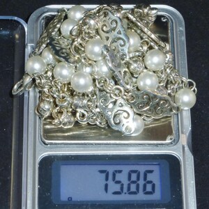 May include: A silver chain necklace with white pearls and ornate silver charms. The necklace is on a digital scale that reads 75.86.